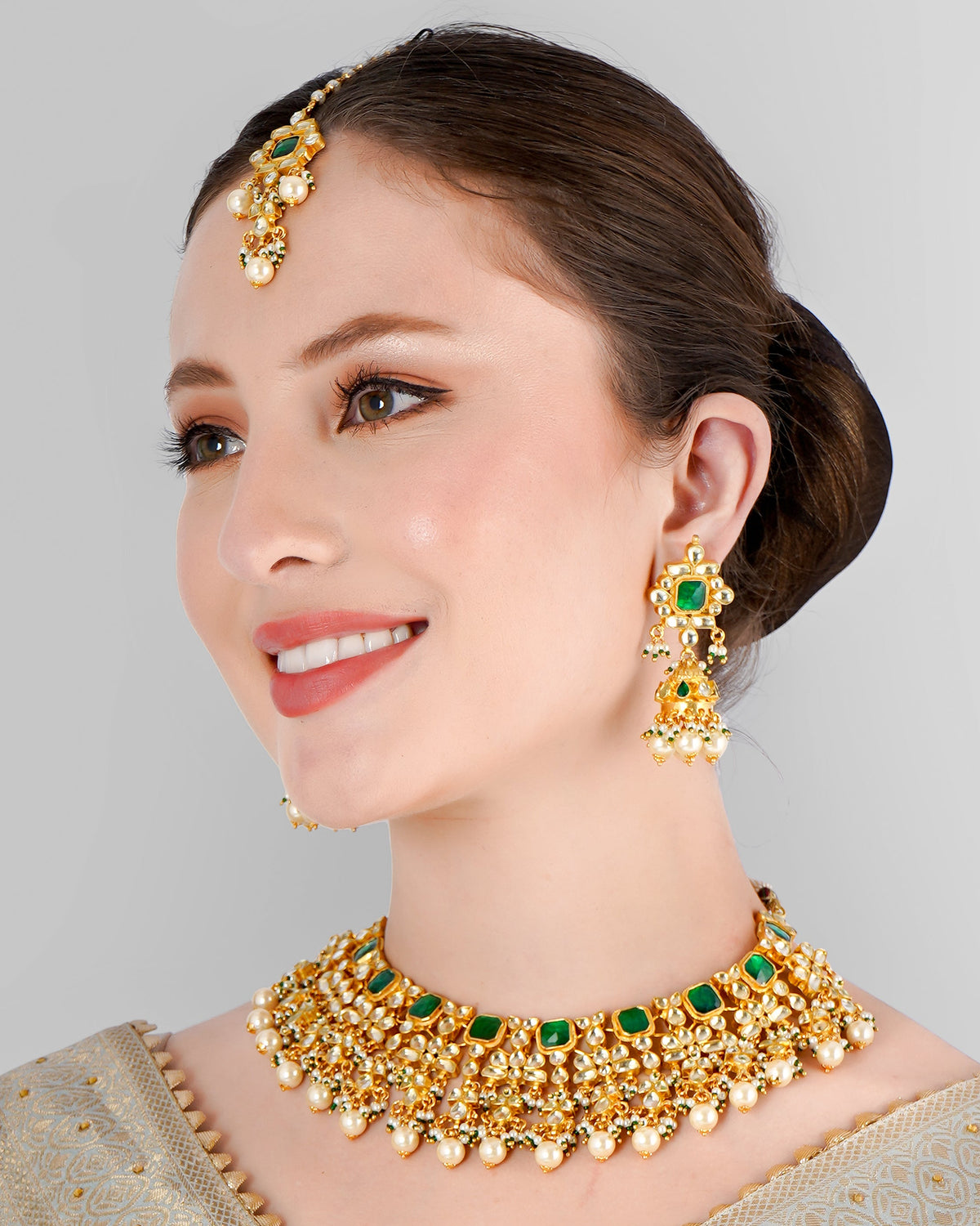A woman wearing a semi-bridal necklace set with green kundan polki and pearls, along with matching earrings and a maangtikka, against a light blue traditional attire.