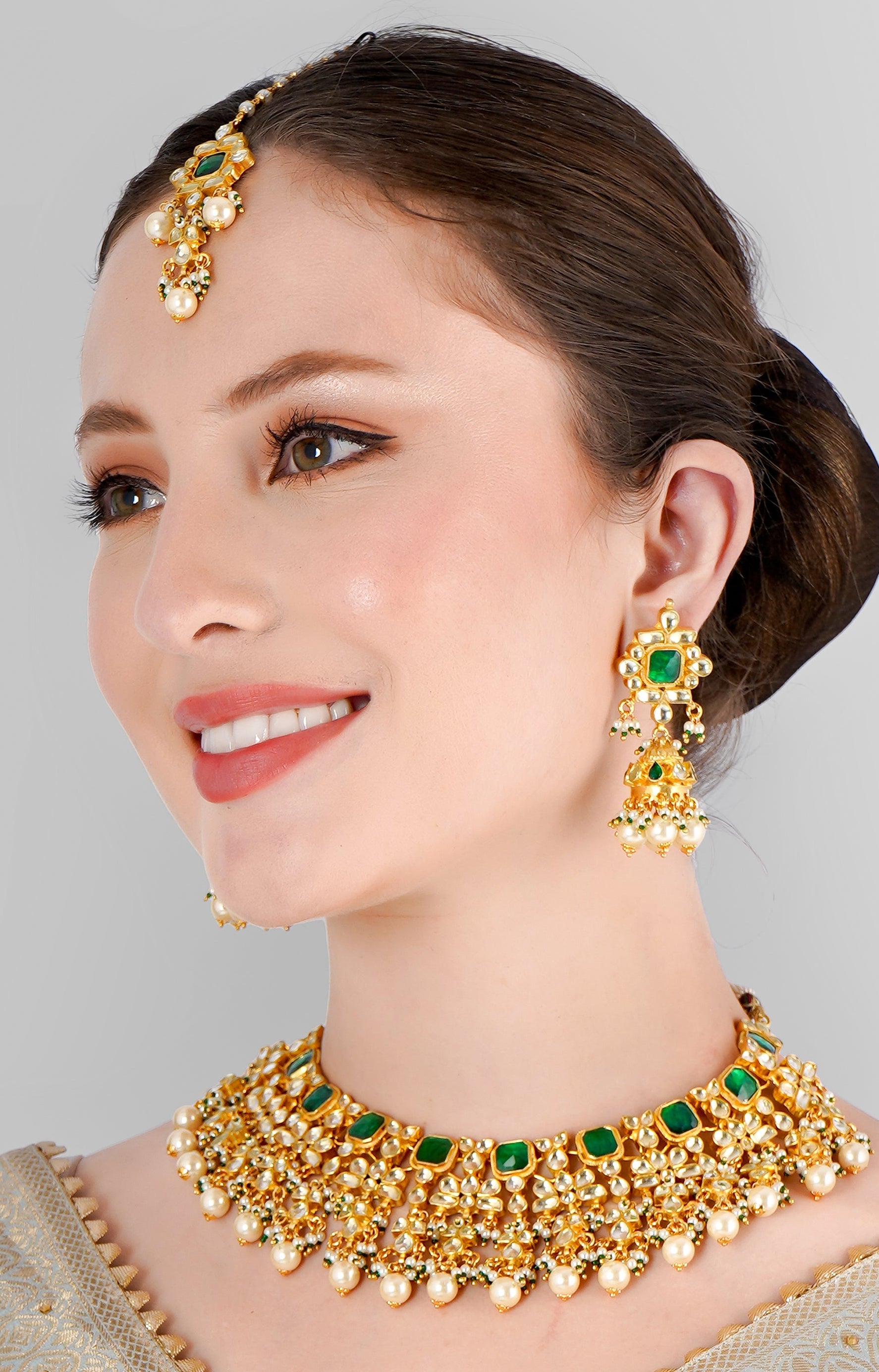 A woman wearing a semi-bridal necklace set with green kundan polki and pearls, along with matching earrings and a maangtikka, against a light blue traditional attire.