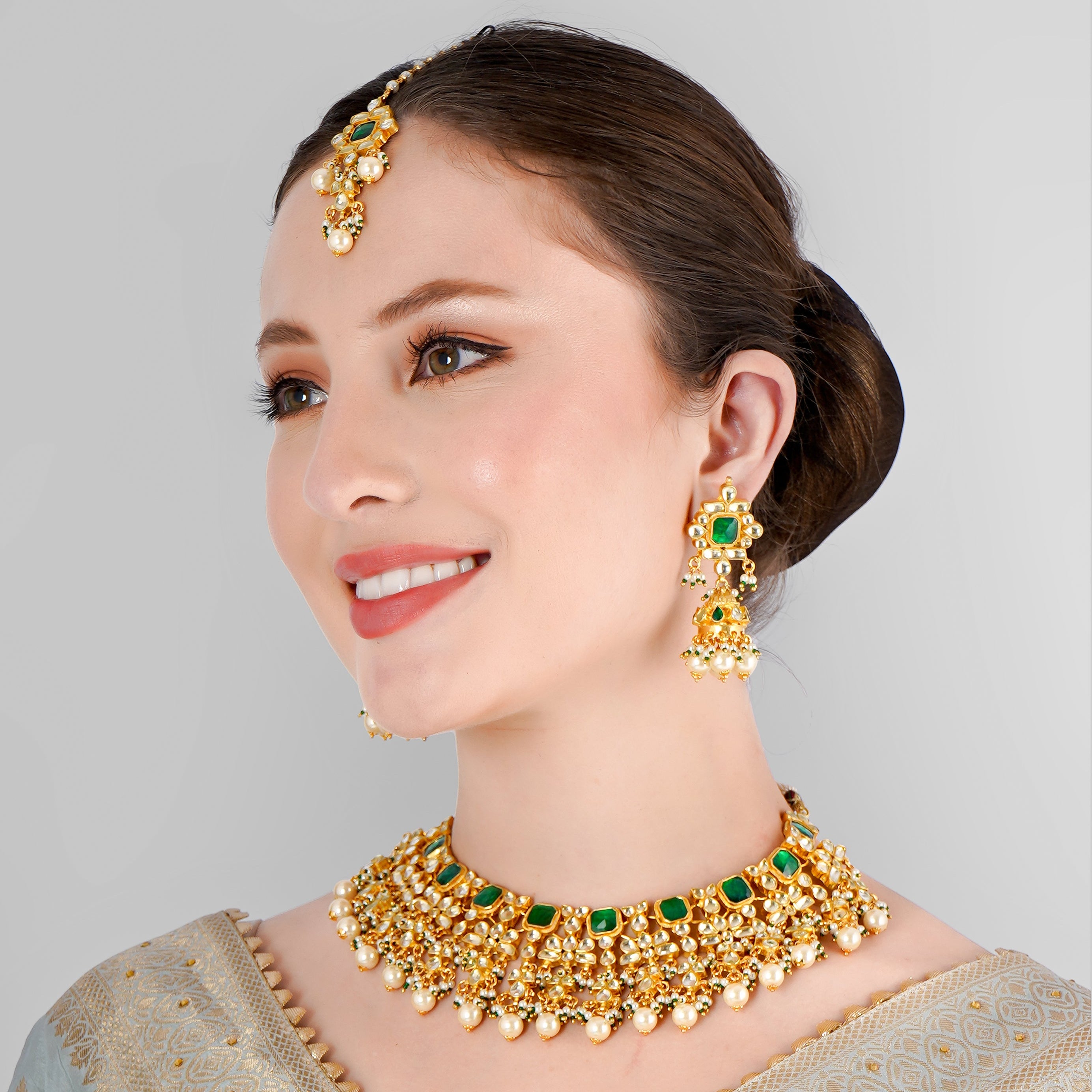 A woman wearing a semi-bridal necklace set with green kundan polki and pearls, along with matching earrings and a maangtikka, against a light blue traditional attire.